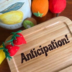 May include: A wooden serving tray with the words "Anticipation!" engraved on the surface. Two red felt strawberries with green leaves are on the tray.