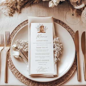May include: A wedding table setting with a menu card featuring a cowboy boot illustration. The menu card is on a white plate with gold silverware, a beige napkin, and floral decorations. The overall theme is rustic and elegant.