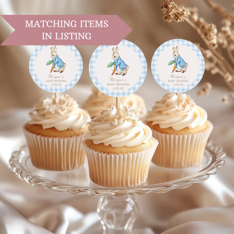 May include: Three white cupcakes with white frosting and blue and white checkered round toppers featuring a cartoon rabbit holding a carrot. The toppers say "Morgan's Baby Shower 4.25.23".