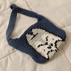 May include: A blue crocheted tote bag with a white crocheted cat face design.