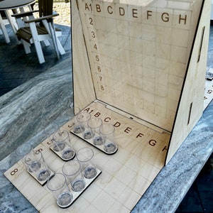 Puede incluir: Un tablero de juego de madera con una cuadrícula de casillas etiquetadas de A1 a H8. El tablero está configurado para un juego de beer pong, con ocho vasos de plástico transparente dispuestos en línea en el tablero.