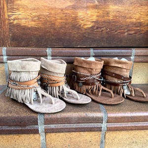 Op de afbeelding: Vier paar su&egrave;de sandalen met franjes in verschillende bruin- en beigetinten. Elke sandaal heeft een teenslipper, leren bandjes en decoratieve accenten. De sandalen worden getoond op een bakstenen trap met een houten achtergrond.