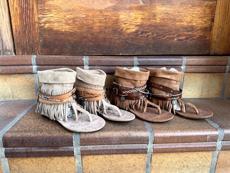 Op de afbeelding: Vier paar su&egrave;de sandalen met franjes in verschillende bruin- en beigetinten. Elke sandaal heeft een teenslipper, leren bandjes en decoratieve accenten. De sandalen worden getoond op een bakstenen trap met een houten achtergrond.