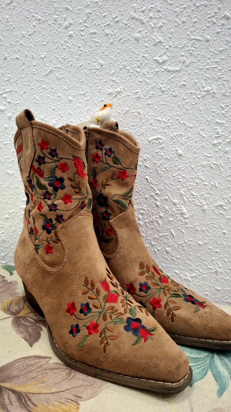 May include: A pair of brown suede ankle boots with embroidered floral designs in red, blue, and green. The boots have a pointed toe and a small heel. A small decorative bird sits on top of the boots.
