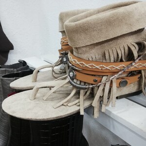 Op de afbeelding: Beige su&egrave;de sandalen met franjes met een bijpassende su&egrave;de laars. De sandalen hebben een teenslipper-ontwerp en zijn versierd met leren bandjes en decoratieve metalen accenten. De laars heeft een omgeslagen manchet en is versierd met leren bandjes en metalen studs.