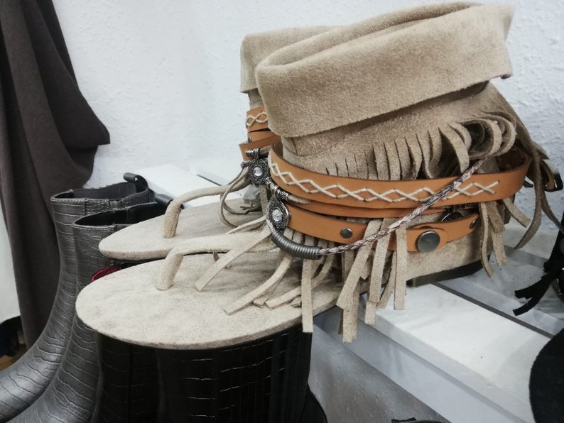 Op de afbeelding: Beige su&egrave;de sandalen met franjes met een bijpassende su&egrave;de laars. De sandalen hebben een teenslipper-ontwerp en zijn versierd met leren bandjes en decoratieve metalen accenten. De laars heeft een omgeslagen manchet en is versierd met leren bandjes en metalen studs.