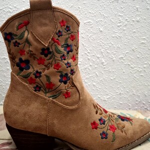 May include: A tan suede ankle boot with a pointed toe and a stacked heel. The boot is embroidered with a floral pattern in red, blue, and green thread. The boot has a pull tab at the top.