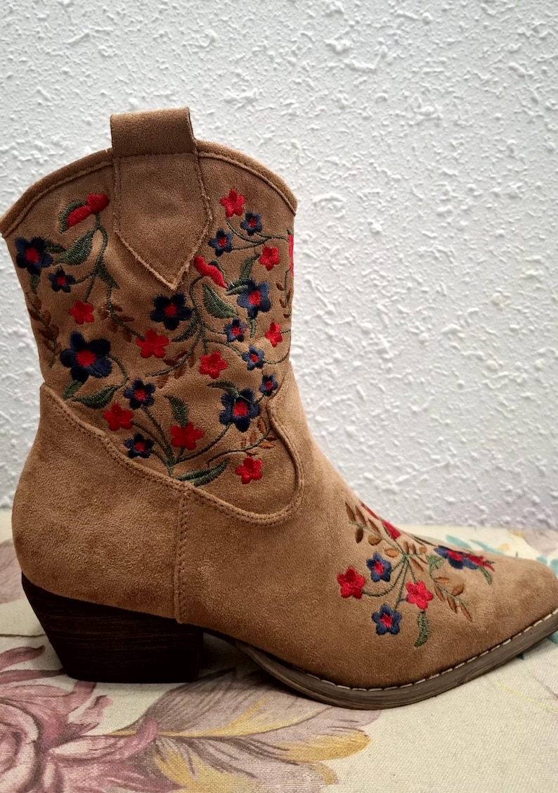 May include: A tan suede ankle boot with a pointed toe and a stacked heel. The boot is embroidered with a floral pattern in red, blue, and green thread. The boot has a pull tab at the top.