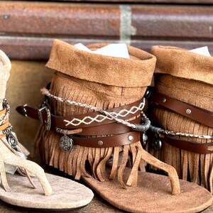 Op de afbeelding: Close-up van bruin leren sandalen met franjes en decoratieve bandjes. De sandalen hebben een teenstijl en een wikkelontwerp rond de enkel. De sandalen worden weergegeven op een bakstenen oppervlak.