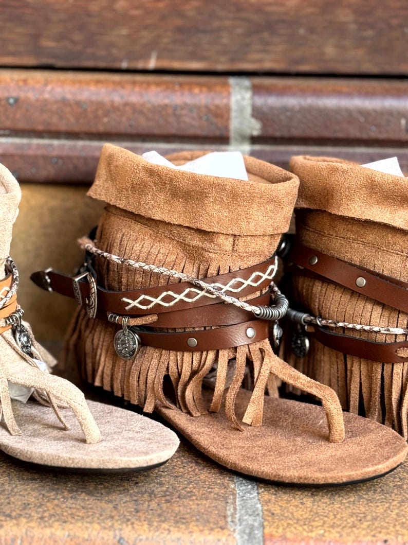 Op de afbeelding: Close-up van bruin leren sandalen met franjes en decoratieve bandjes. De sandalen hebben een teenstijl en een wikkelontwerp rond de enkel. De sandalen worden weergegeven op een bakstenen oppervlak.