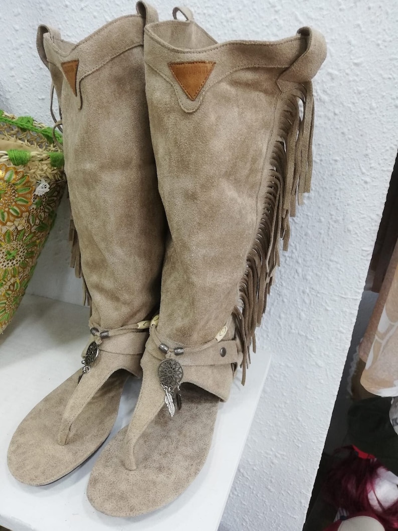 May include: A pair of beige suede leather boots with a thong sandal design. The boots feature fringe detailing along the sides, decorative metal charms, and a small leather triangle patch at the top.