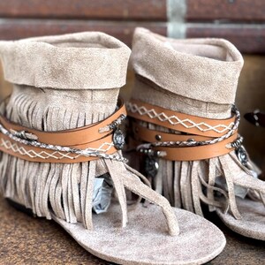 Op de afbeelding: Een paar beige su&egrave;de franje sandalen met bruine leren bandjes en zilverkleurige versieringen. De sandalen hebben een teenstuk in teenslipperstijl en een omgeslagen enkelmanchet. Het schoeisel wordt gepresenteerd op een bakstenen oppervlak.