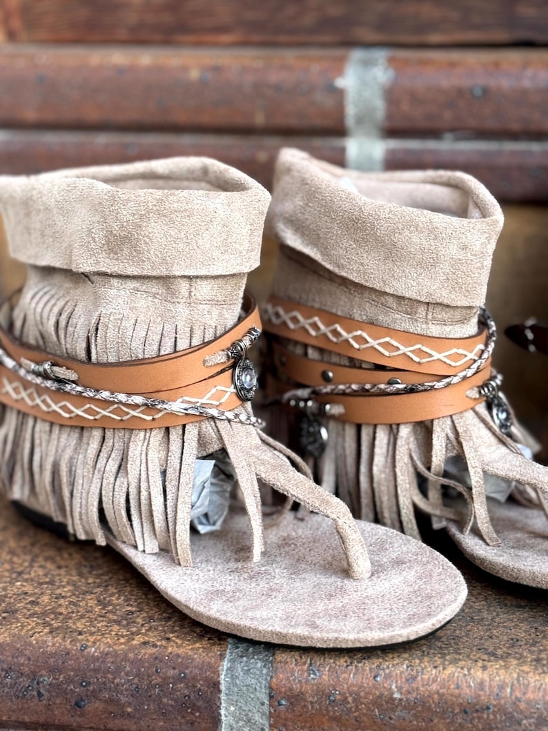 Op de afbeelding: Een paar beige su&egrave;de franje sandalen met bruine leren bandjes en zilverkleurige versieringen. De sandalen hebben een teenstuk in teenslipperstijl en een omgeslagen enkelmanchet. Het schoeisel wordt gepresenteerd op een bakstenen oppervlak.