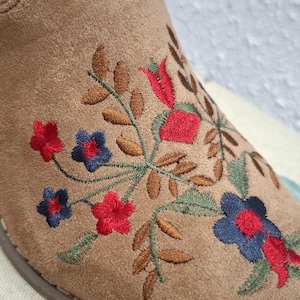 May include: Close-up of a tan suede boot with floral embroidery. The embroidery features red, blue, and green flowers and leaves. The boot has a rounded toe and a brown sole. The overall style is bohemian.