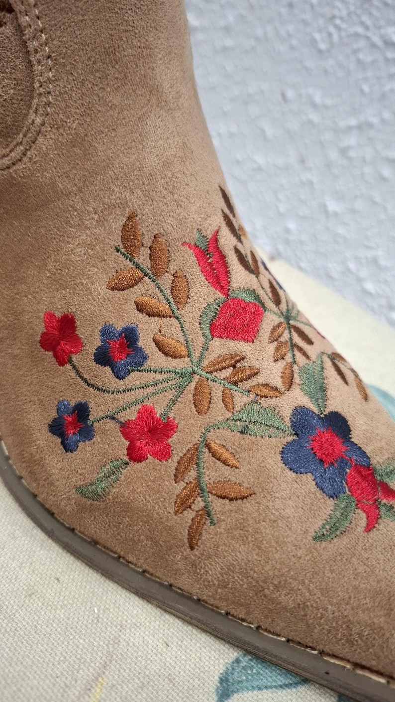 May include: Close-up of a tan suede boot with floral embroidery. The embroidery features red, blue, and green flowers and leaves. The boot has a rounded toe and a brown sole. The overall style is bohemian.