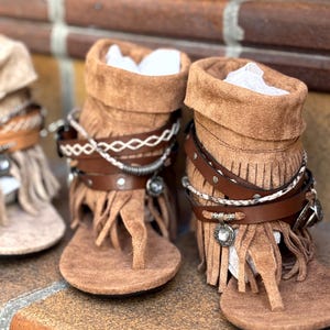 Op de afbeelding: Bruine su&egrave;de sandalen met franjes en decoratieve bandjes. De sandalen hebben een teen van het teenslipper-type en een kort laarsachtig ontwerp. De bandjes bevatten zilverkleurige versieringen en een leren band. De sandalen worden getoond op een bakstenen oppervlak.