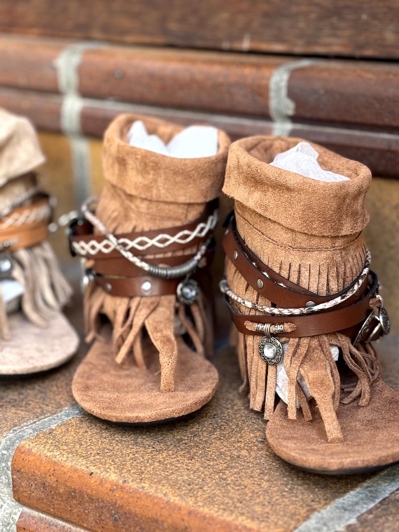 Op de afbeelding: Bruine su&egrave;de sandalen met franjes en decoratieve bandjes. De sandalen hebben een teen van het teenslipper-type en een kort laarsachtig ontwerp. De bandjes bevatten zilverkleurige versieringen en een leren band. De sandalen worden getoond op een bakstenen oppervlak.