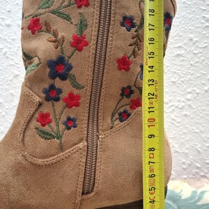 May include: Tan suede ankle boot with embroidered red and blue flowers. The boot has a side zipper and a stacked heel. The boot's height is approximately 10 inches.