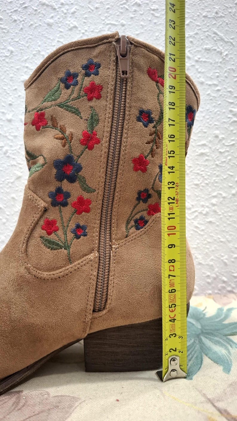 May include: Tan suede ankle boot with embroidered red and blue flowers. The boot has a side zipper and a stacked heel. The boot's height is approximately 10 inches.