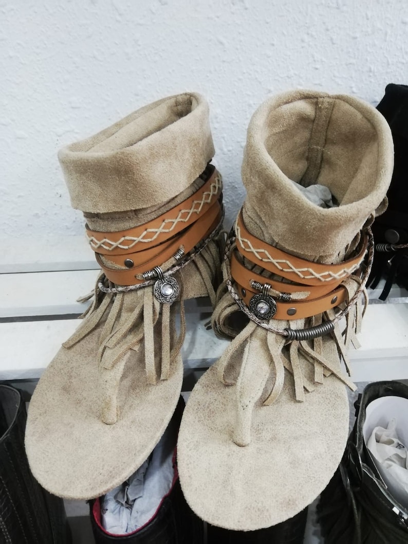 Op de afbeelding: Een paar beige su&egrave;de sandalen met een teenslipper ontwerp. De sandalen hebben een gefranjerde teen, leren bandjes en metalen accenten. De enkelmanchetten zijn omgeslagen. De sandalen worden op een witte plank getoond. Ongeveer 25 cm lang.