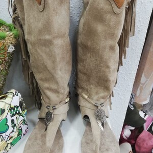 May include: A pair of beige suede thong sandals with knee-high shafts. The boots have decorative fringe, metal charms, and a small leather patch. The sandals are displayed on a white surface.