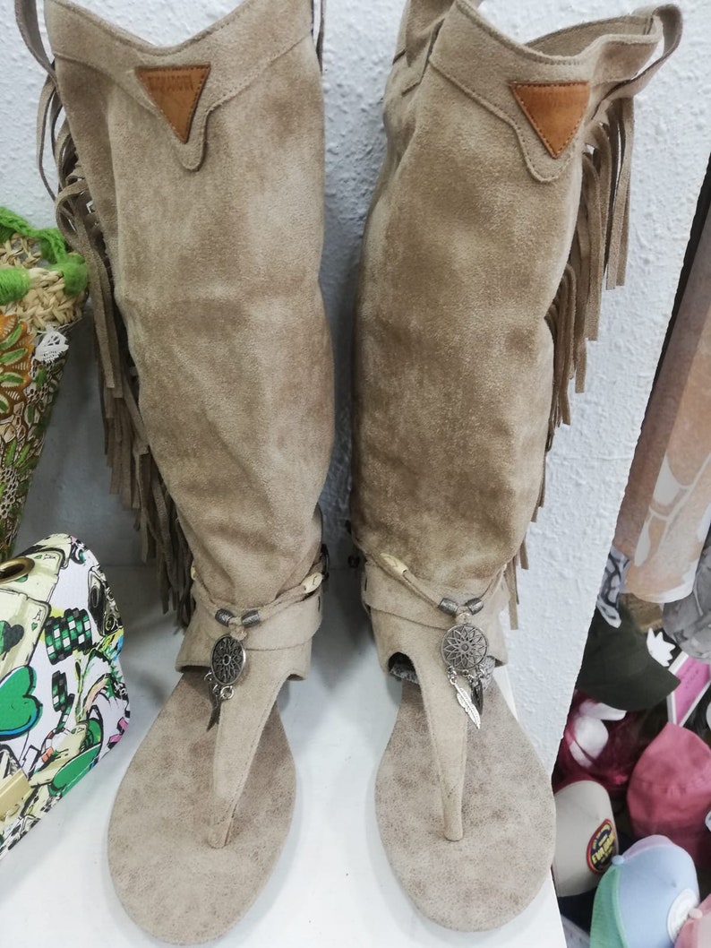 May include: A pair of beige suede thong sandals with knee-high shafts. The boots have decorative fringe, metal charms, and a small leather patch. The sandals are displayed on a white surface.