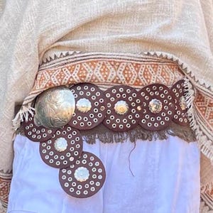 Op de afbeelding: Een bruine leren riem met zilveren versieringen en een grote zilveren gesp. De riem heeft cirkelvormige uitsparingen en decoratieve klinknagels. De riem wordt gedragen over een witte short, met een gefranjerde rand en een patroonstof op de achtergrond.