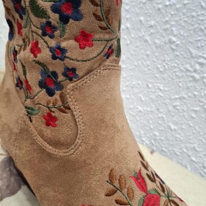 May include: Close-up of a tan suede boot with embroidered floral detailing. The embroidery features red, blue, and green flowers and leaves. The boot has a classic western style.