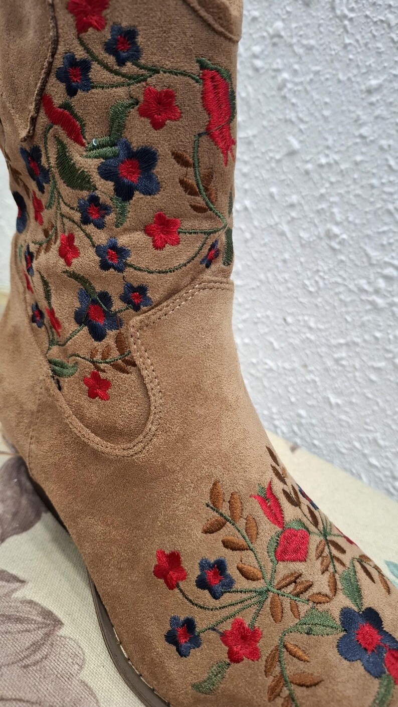May include: Close-up of a tan suede boot with embroidered floral detailing. The embroidery features red, blue, and green flowers and leaves. The boot has a classic western style.