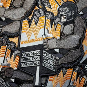 Op de afbeelding: Een stapel geborduurde patches met een gorilla die een Whataburger-beker vasthoudt en een bord met de tekst "Open 24 Hrs Drive Thru Breakfast". De patches zijn zwart-wit met gele en oranje accenten.