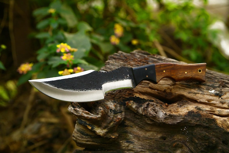 Puede incluir: Un cuchillo de caza negro y plateado con mango de madera. El cuchillo est&aacute; apoyado en un trozo de madera desgastada.
