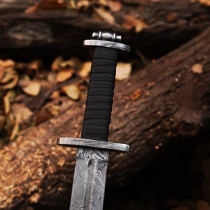 May include: A close-up of the handle of a black-handled sword with a Damascus steel blade. The sword is resting on a wooden surface.