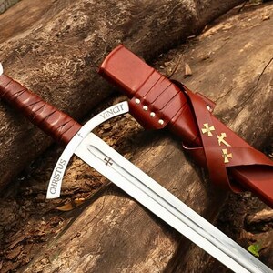 May include: A medieval-style sword with a silver blade and a brown leather-wrapped handle. The sword features the inscription "CHRISTUS VINCIT" and is accompanied by a matching brown leather sheath with gold cross details. The sword rests on a wooden surface.