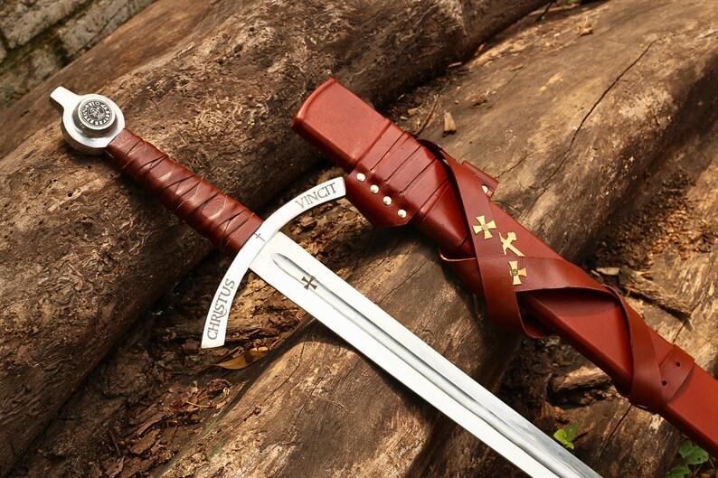 May include: A medieval-style sword with a silver blade and a brown leather-wrapped handle. The sword features the inscription "CHRISTUS VINCIT" and is accompanied by a matching brown leather sheath with gold cross details. The sword rests on a wooden surface.