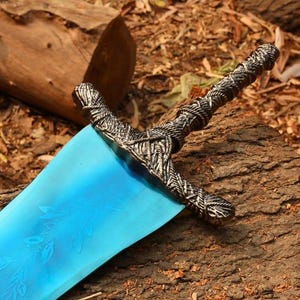 May include: A fantasy sword featuring a vibrant blue blade and a detailed, textured silver-coloured hilt. The blade has a floral design etched into it. The sword rests on a piece of wood, with a natural background.