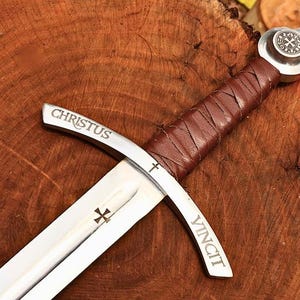 May include: A medieval-style sword with a silver blade and a brown leather-wrapped handle. The crossguard is inscribed with the words "CHRISTUS VINCIT" and a cross symbol. The sword rests on a wooden surface.