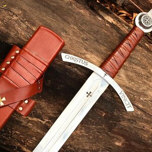 May include: A medieval sword with a brown leather-wrapped handle and a silver blade. The blade has the words "CHRISTUS VINCIT" and a cross symbol. The sword is next to a brown leather sheath with a cross.