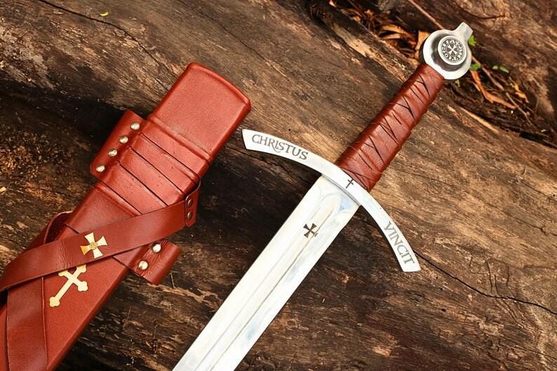 May include: A medieval sword with a brown leather-wrapped handle and a silver blade. The blade has the words "CHRISTUS VINCIT" and a cross symbol. The sword is next to a brown leather sheath with a cross.