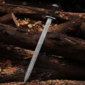 May include: A silver sword with a black leather handle, lying on a pile of brown tree logs.