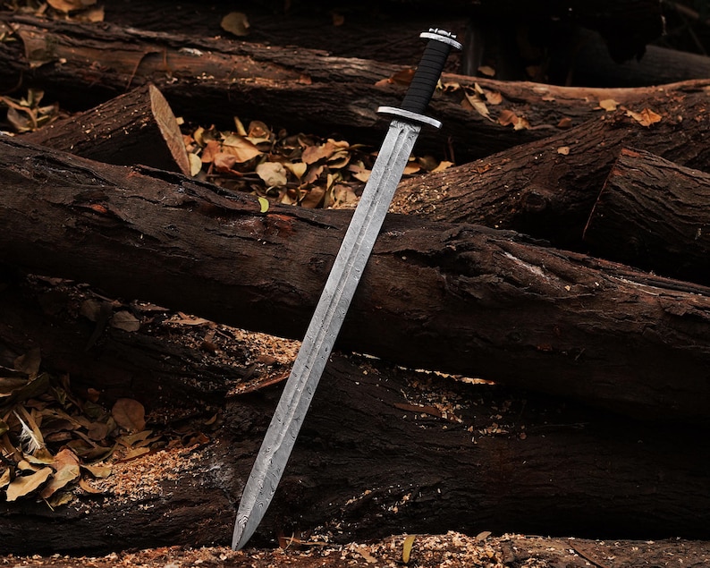 May include: A silver sword with a black leather handle, lying on a pile of brown tree logs.