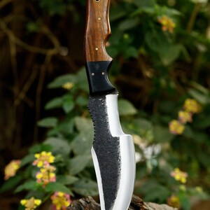 Puede incluir: Un cuchillo de caza negro y plateado con mango de madera. El cuchillo est&aacute; sobre un trozo de madera con un fondo verde borroso.