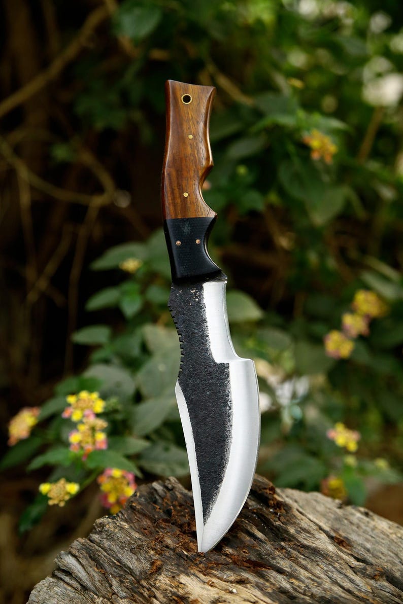 Puede incluir: Un cuchillo de caza negro y plateado con mango de madera. El cuchillo est&aacute; sobre un trozo de madera con un fondo verde borroso.