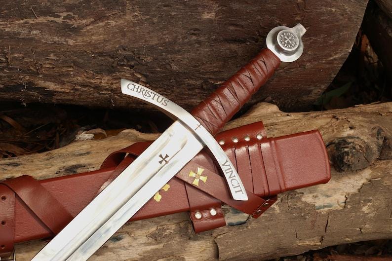 May include: A medieval-style sword with a silver blade and a brown leather-wrapped handle. The sword is resting on a brown leather sheath. The blade has the words "CHRISTUS" and "VINCIT" etched on it, along with a cross symbol.