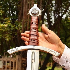 May include: A medieval-style sword with a silver blade and a brown leather-wrapped handle. The crossguard is inscribed with the words "CHRISTUS VINCIT". The sword has a silver pommel with a circular emblem.