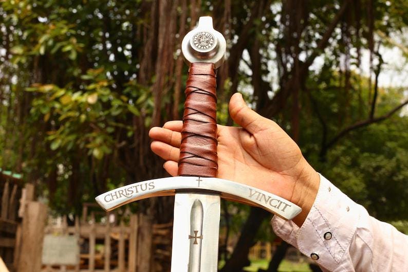 May include: A medieval-style sword with a silver blade and a brown leather-wrapped handle. The crossguard is inscribed with the words "CHRISTUS VINCIT". The sword has a silver pommel with a circular emblem.