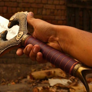 May include: A sword featuring a metallic blade with etched symbols, a brass-colored hilt shaped like a bird head with intricate scale detailing, and a dark brown handle wrapped with thin cord, providing a firm grip for the user holding the weapon.