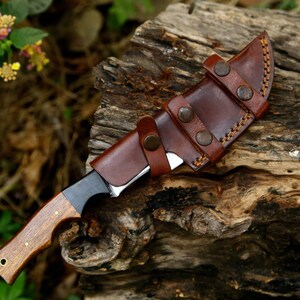 Puede incluir: Una funda de cuero marr&oacute;n con costuras y broches met&aacute;licos contiene un cuchillo con mango negro y mango de madera. El cuchillo y la funda est&aacute;n apoyados sobre un trozo de madera desgastada.