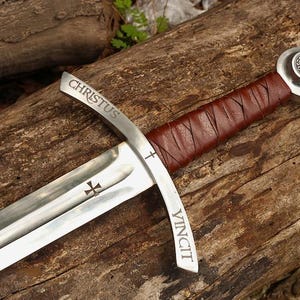 May include: A medieval sword with a silver blade and a brown leather-wrapped handle. The blade has the words "CHRISTUS VINCIT" etched on it, along with a cross symbol. The sword rests on a wooden surface.