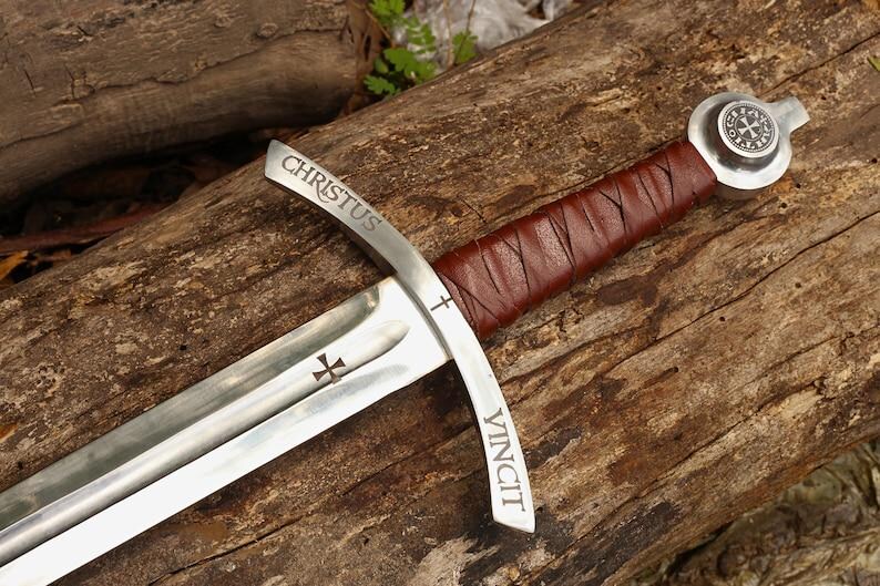 May include: A medieval sword with a silver blade and a brown leather-wrapped handle. The blade has the words "CHRISTUS VINCIT" etched on it, along with a cross symbol. The sword rests on a wooden surface.