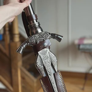 Pode incluir: Um close-up detalhado de uma espada prateada e castanha. A espada apresenta desenhos intrincados na guarda e na lâmina, com uma pega e detalhes em couro castanho. A lâmina tem um padrão decorativo. A espada é segurada por uma mão.